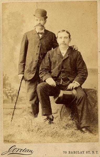 Sepia portrait photograph of two men, wearing suits, one seated with a top hat, one standing with a bowler hat and cane, in the studio of Jordan, New York. Taken in the late 19th century, this highlights the popularity of the bowler hat. Newry and Mourne Museum Collection
