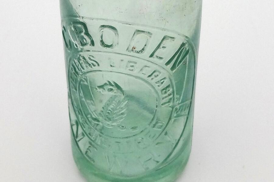 Moulded clear glass bottle, slightly wider at the shoulder than the base, long neck, would have had a metal bottlecap, label worked in glass 'C. Boden, Newry, registered trademark, Veritas Liberabit', 'wholesale bottlers' around base, 'British made, K. & Co. B., M586' on base. Newry & Mourne Museum Collection