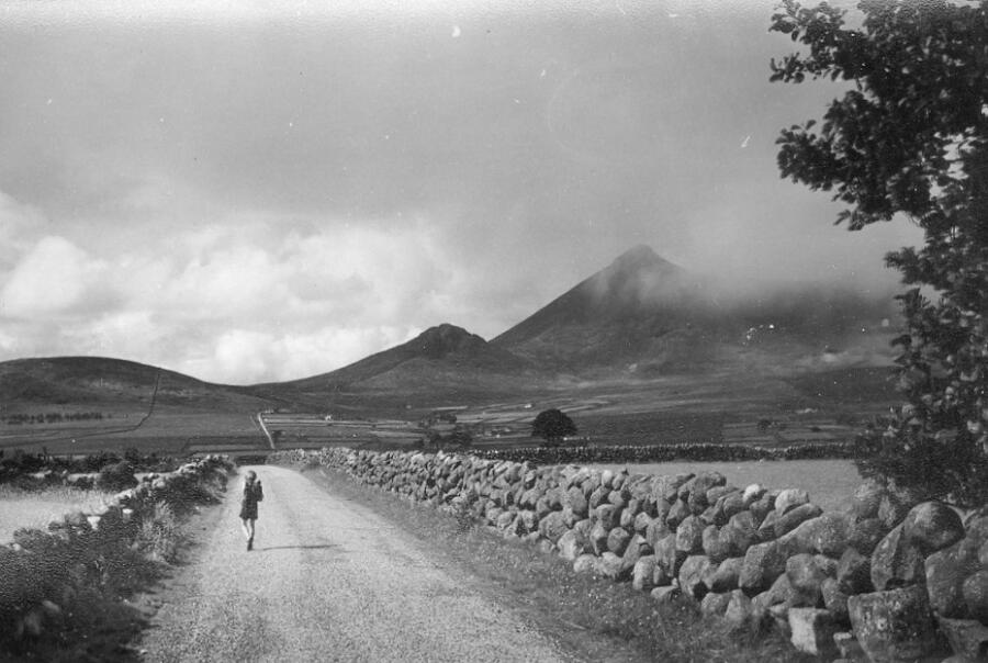 Black and white photograph of a school child walking along a road, and stone walls in the Mourne Mountains, 1960s. Newry and Mourne Museum Collection